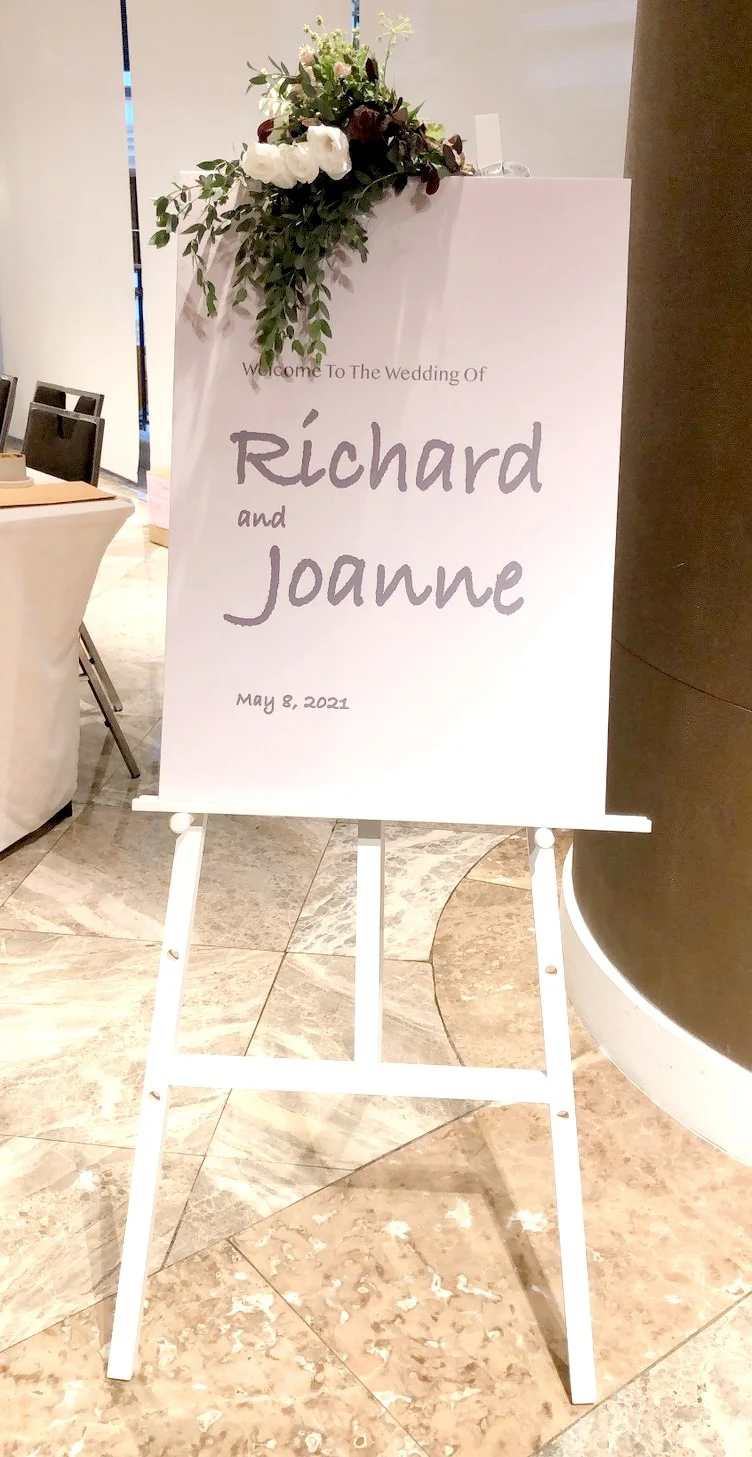 白色立架上放置婚禮歡迎牌,牌面寫著Richard和Joanne的婚禮日期2021年5月8日,牌頂裝飾白色、深紅色花朵及綠色垂掛葉片