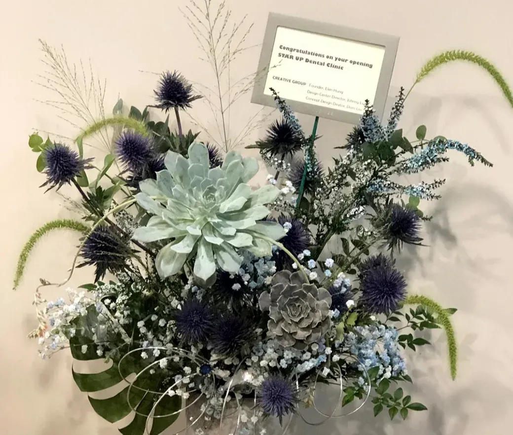 白色背景前放置一個花卉插花作品,含淺綠色多肉植物、深紫色球狀花朵、淺藍色小花、細長綠色草狀植物及白色小花,背後豎立一張祝賀卡片