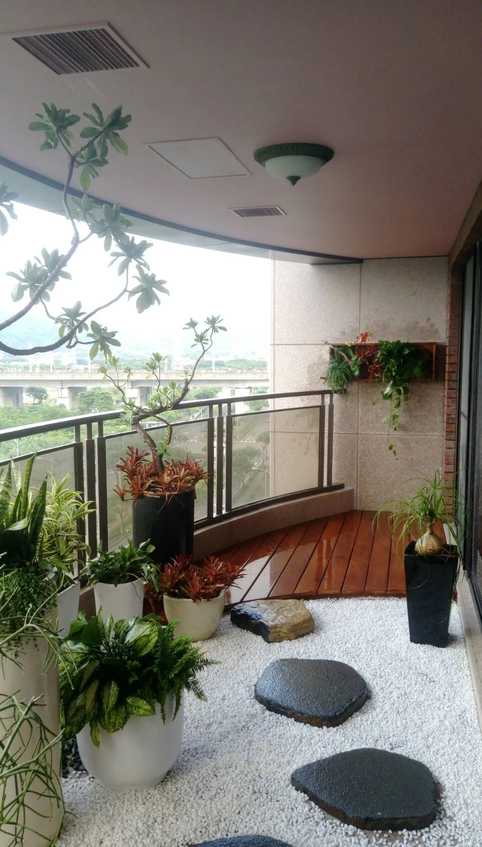 高樓陽台上擺放多個綠色盆栽與紅色花卉,木質地板與白色石子地面,玻璃護欄外可見橋樑與遠方景觀