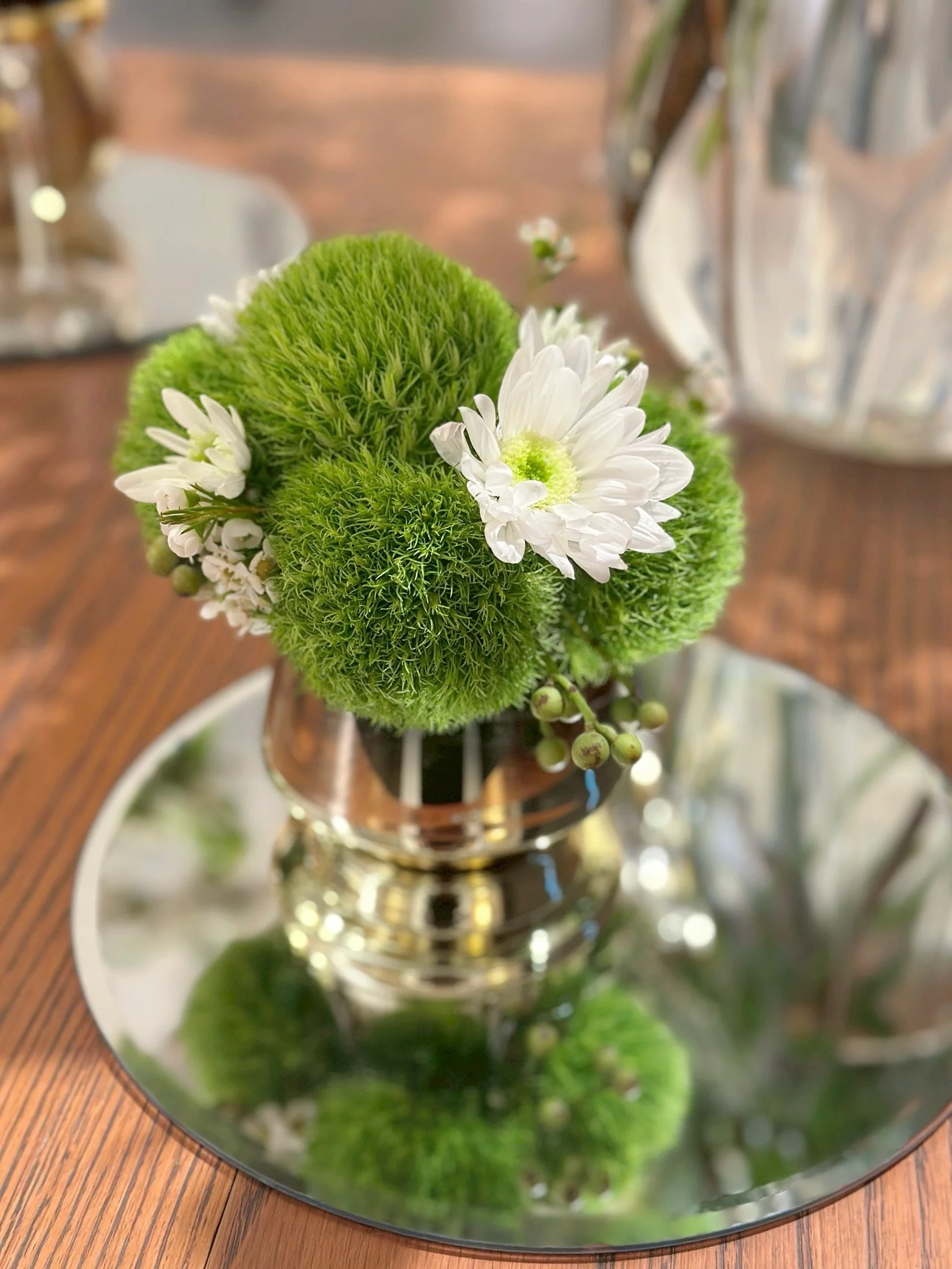 玻璃花瓶內插著亮綠色球狀苔蘚、白色小花和綠色莖葉,放置於木紋桌面上的圓形玻璃墊