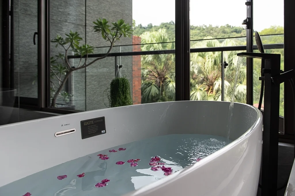 白色浴缸內盛滿淡藍色水,散放紅紫色花瓣,背景為落地窗和綠色植被景觀