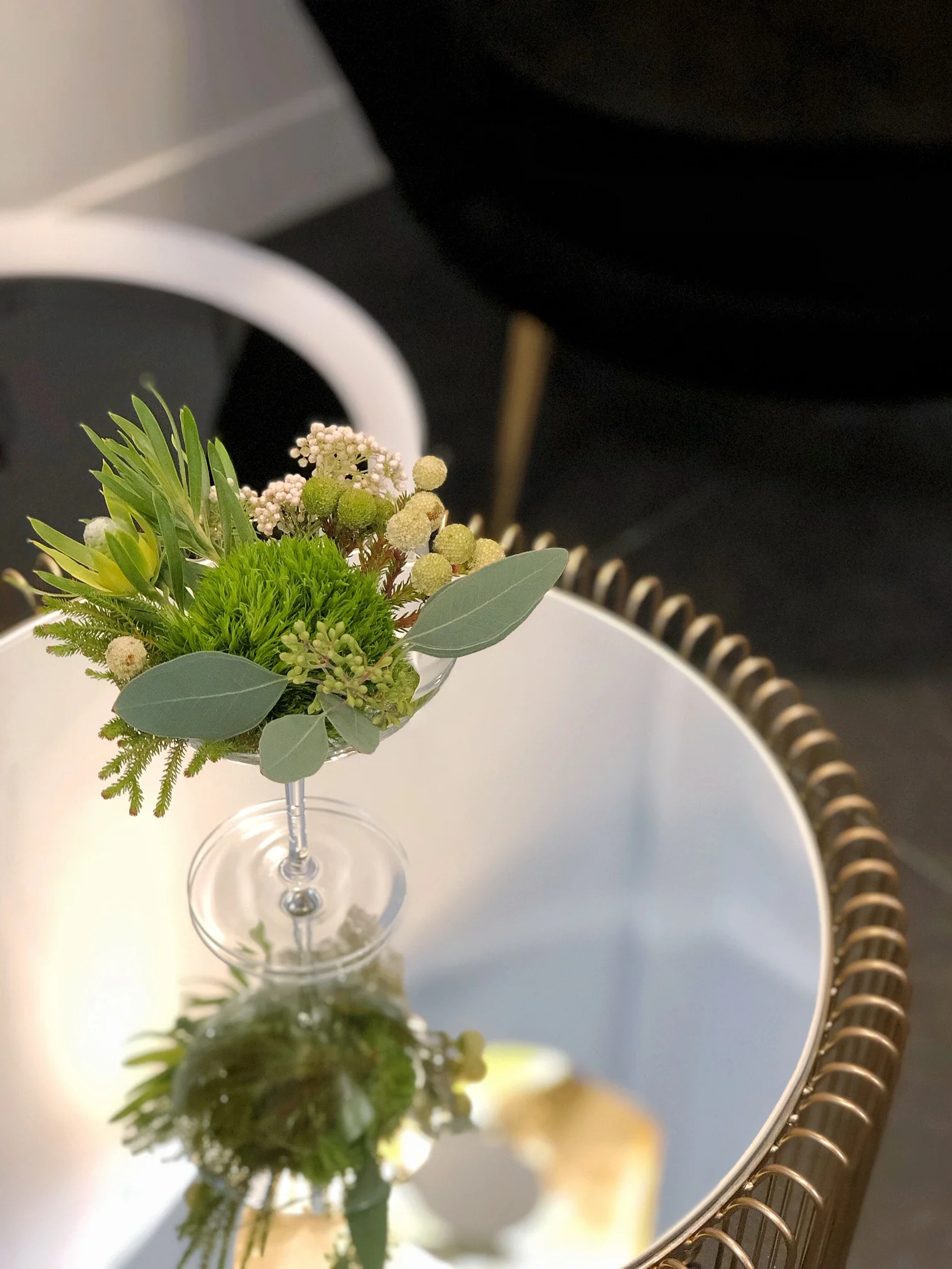 玻璃花瓶中插著綠色葉片、白色小花和球形花蕾的花藝作品,背景為白色圓形鏡子和金色邊框裝飾