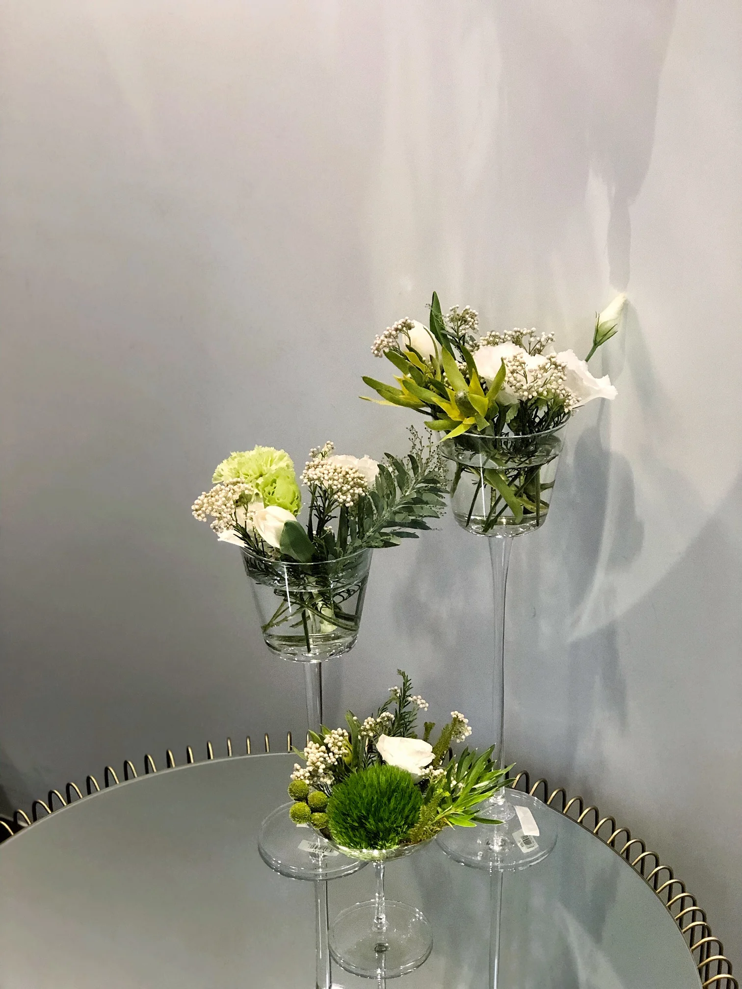白色背景前,玻璃杯中插著白色和黃綠色花朵、細小白色小花及綠葉,三個花瓶高度遞增排列於圓形金邊盤上