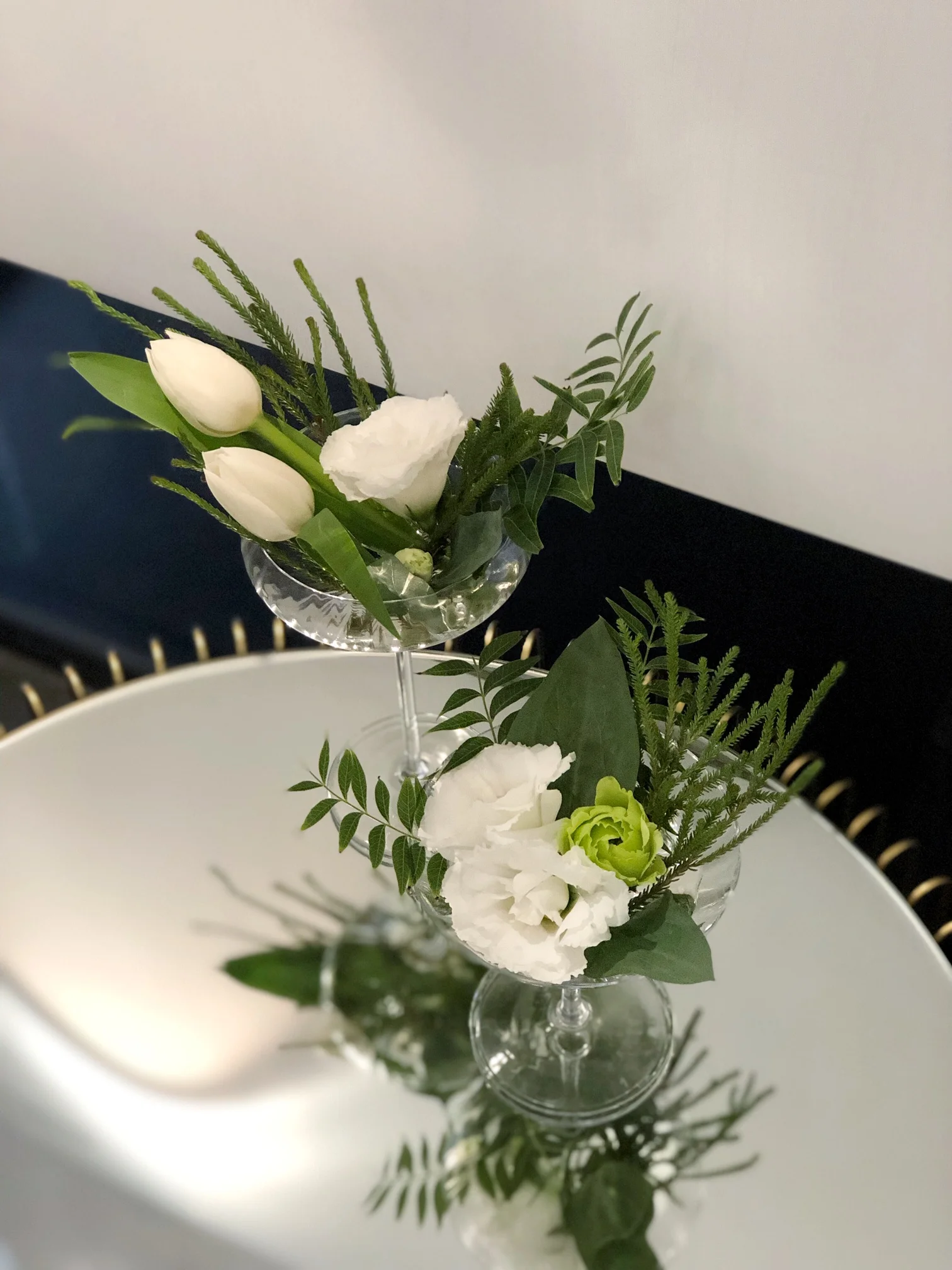 兩個透明玻璃杯中插著白色花朵與綠色葉材,放在白色盤子旁,背景為灰白色牆面和黑色物體