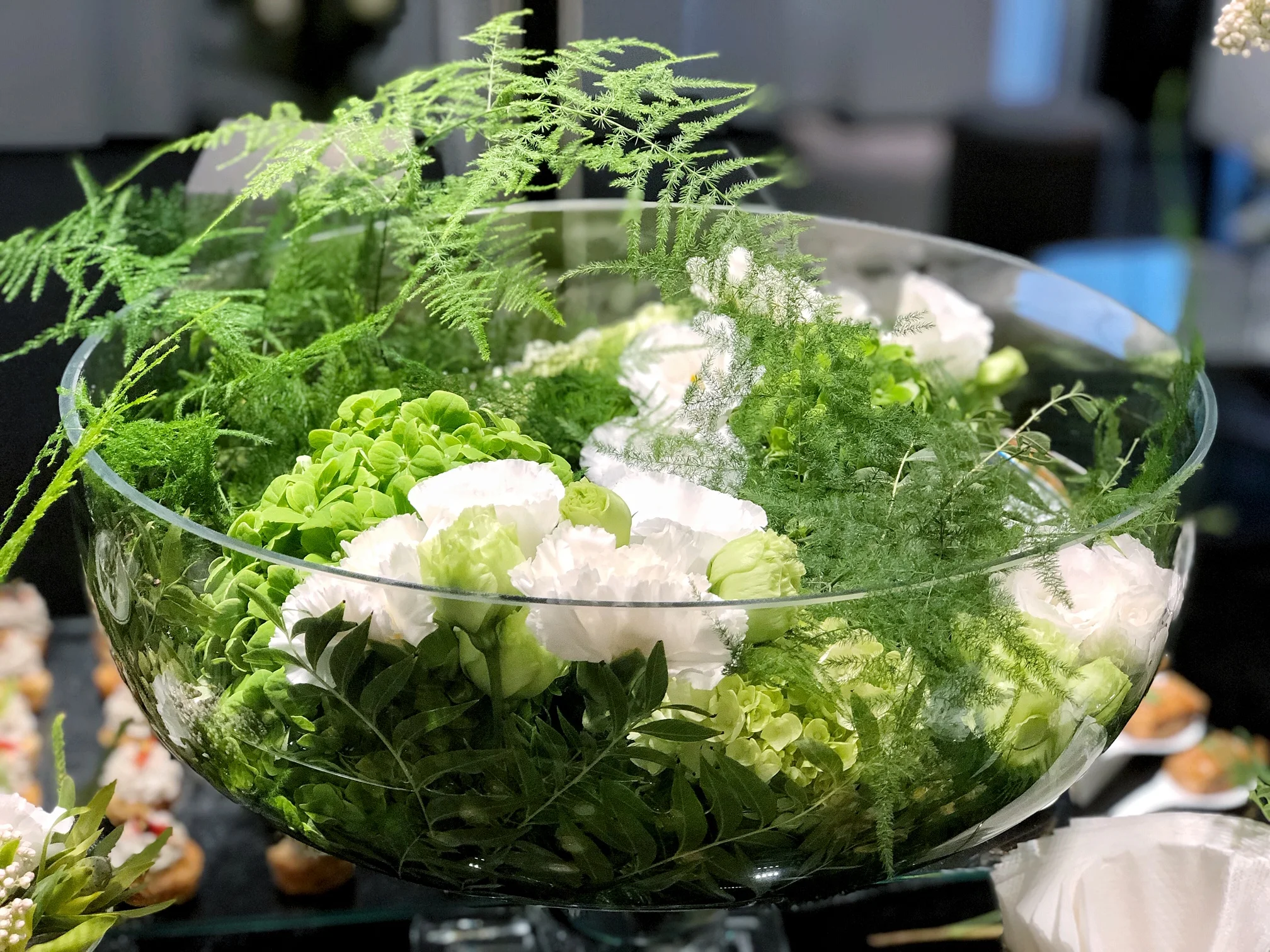 透明玻璃碗內盛放綠色蕨類植物、白色小花、淡綠色花朵和各式綠葉,放置在室內桌面上