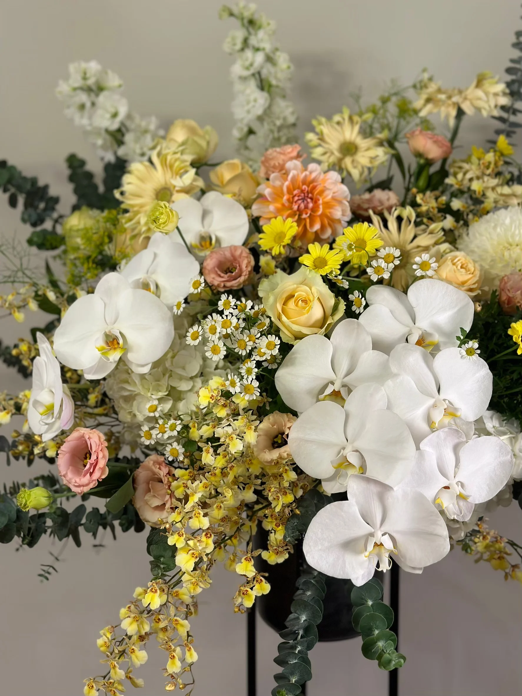 白色蘭花為主體,搭配黃色小花、粉紅玫瑰、橙色大花和白色小雛菊,插在黑色花器中的豐滿花束
