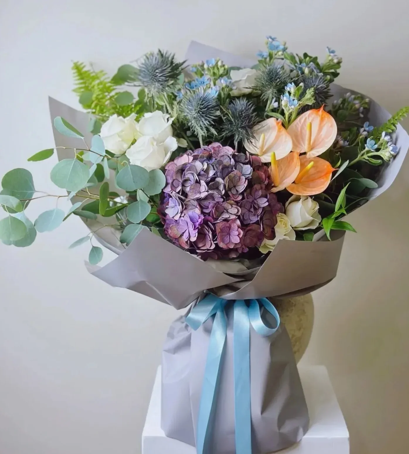 灰色紙質包裝的花束,內含紫色繡球花、粉橙色花朵、白色花卉、藍色小花、綠葉和尤加利葉,下方繫著藍白色緞帶