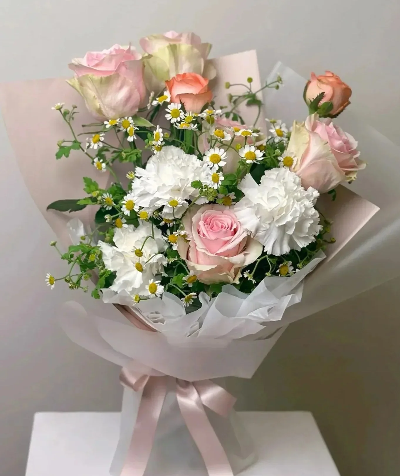 粉紅色玫瑰、白色花卉、黃白小雛菊和綠葉組成的花束,用白色紙張包裝,繫著粉紅色蝴蝶結