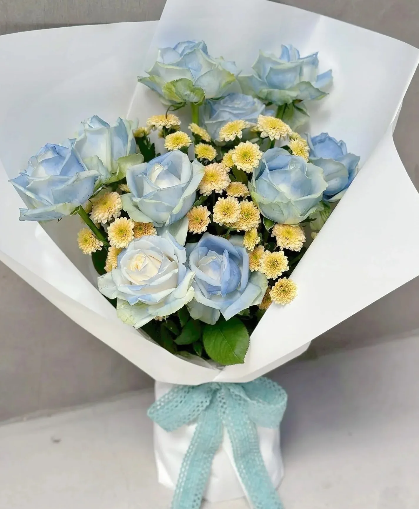白色包裝紙束中,淺藍色玫瑰花和黃色小菊花組成的花束,綁著淺藍色蝴蝶結