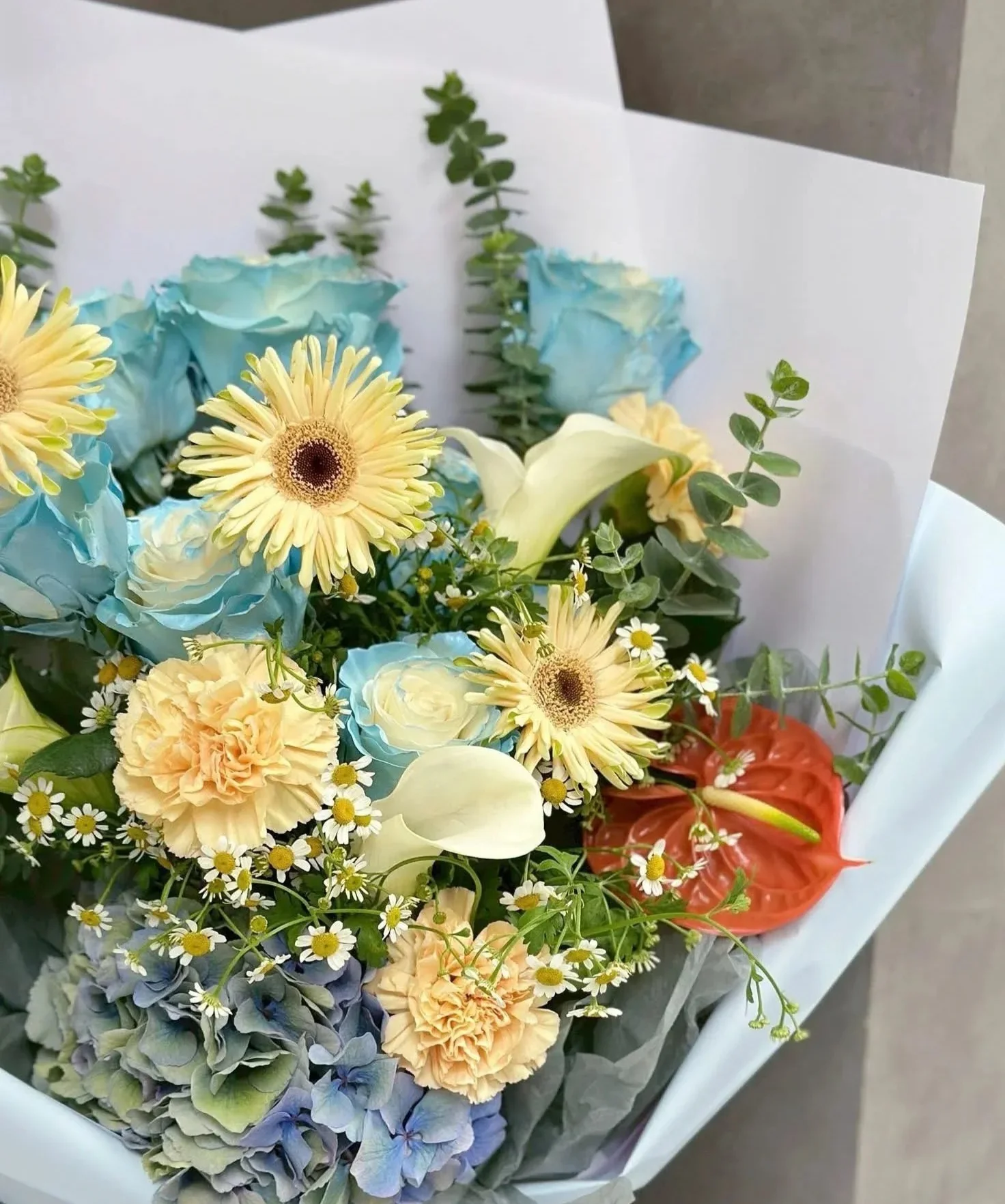 白色紙質包裝的花束,包含黃色菊花、藍色玫瑰、米色康乃馨、白色馬蹄蓮、紅色花朵及綠色葉材與小白花