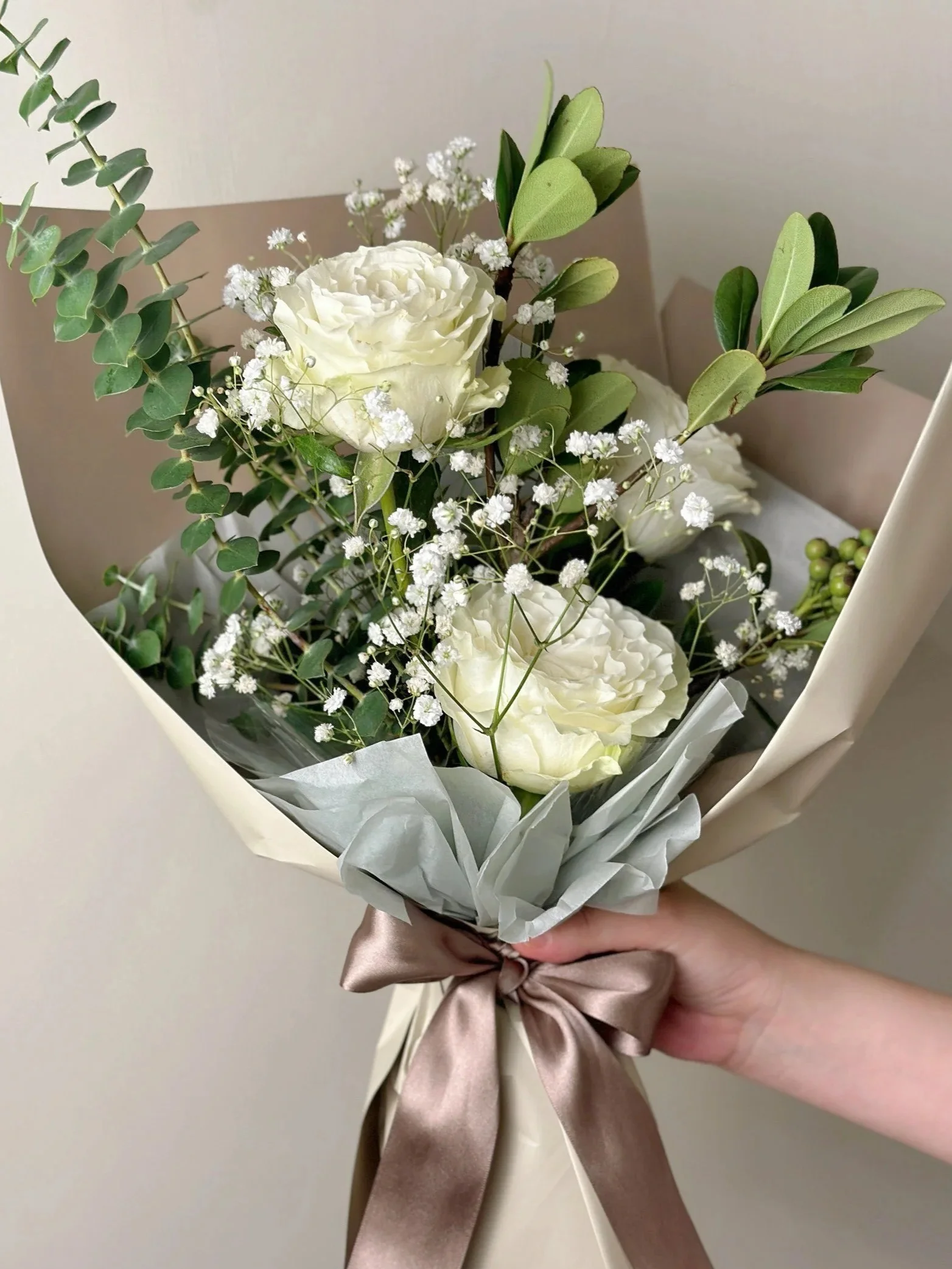 手持一束白色包裝花束,內含兩朵白色玫瑰、白色小花、綠色葉材,以棕色緞帶綁結