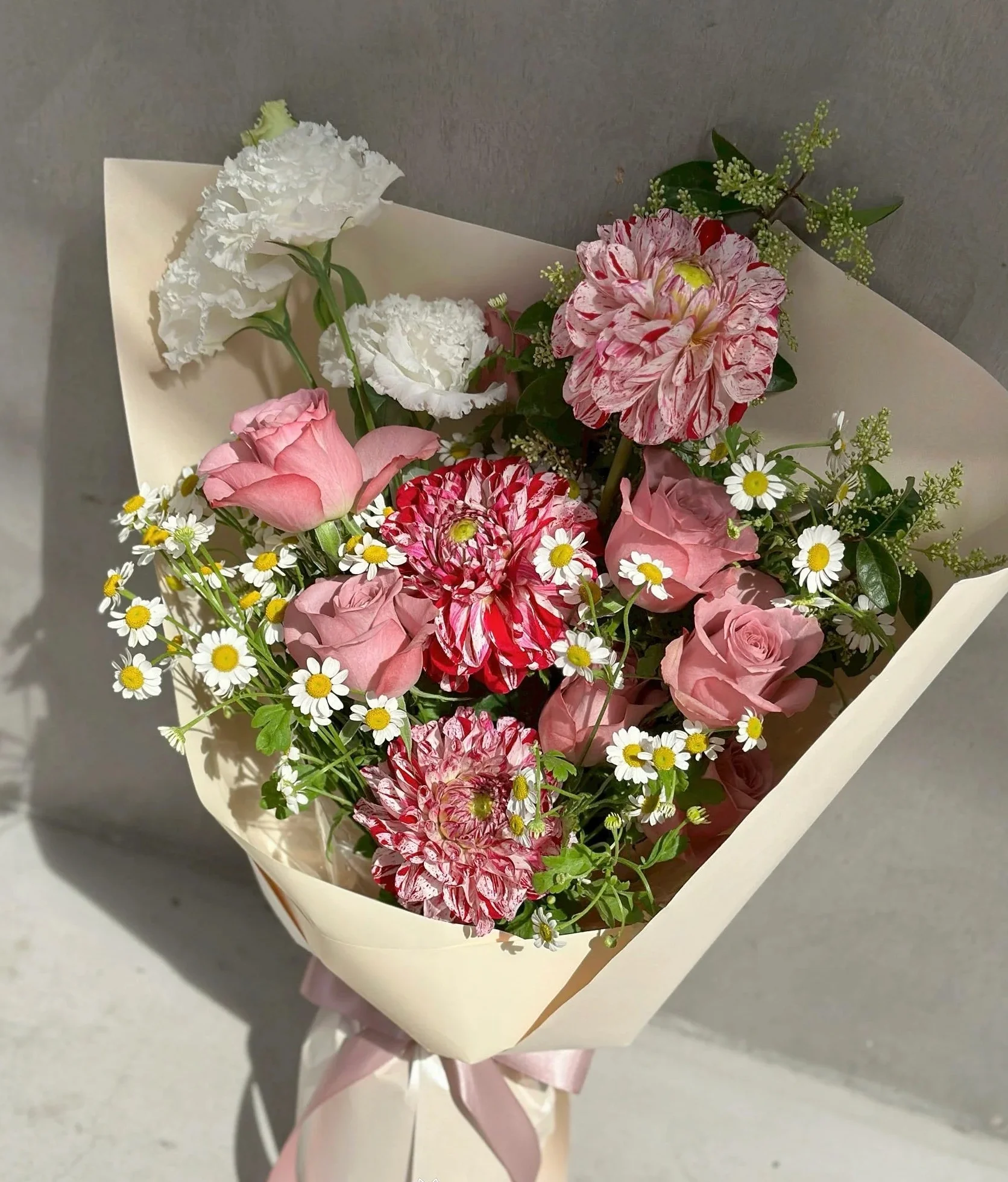 白色包裝紙包裹的花束,內含白色牡丹、粉紅玫瑰、紅白相間牡丹、白色小菊花和綠色裝飾草,配粉紅絲帶